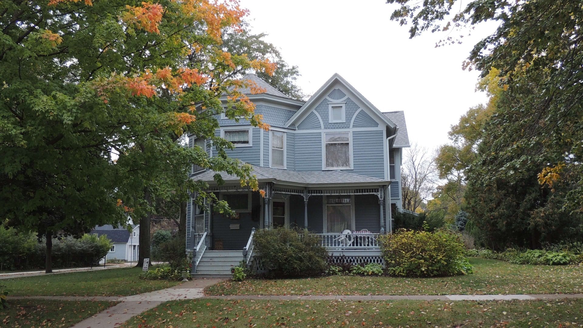 Ash Park 703 6th Ave NW. This is a 2-story Queen Anne home with a large tower and a broad covered porch. The home is blue with white trim. The grounds are pleasantly planted with shrubs and flowers.