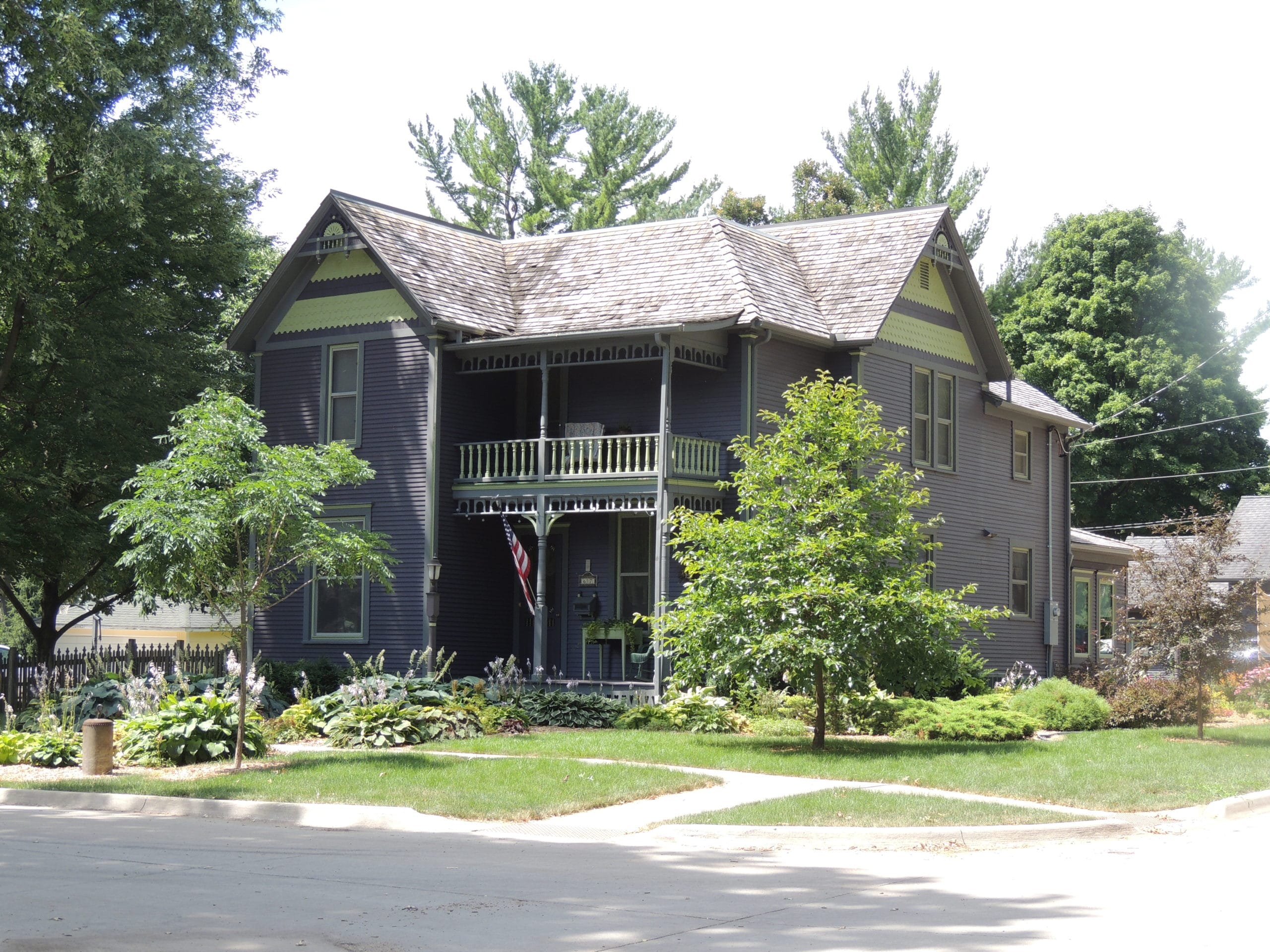617 7th Avenue NW, front side view. A tall 2-story home with dark slate blue/purple siding with gray trim accented with large bargeboard peak decorations and yellow fish scale siding on the roof gables. The front covered porch has a 2nd-story "sleeper porch" above.