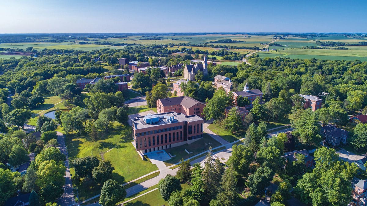 Aerial view of Cornell College campus as seen from the South East. A picturesque treelined hillside with blue sky and many large brick classroom buildings peppered on the landscape.